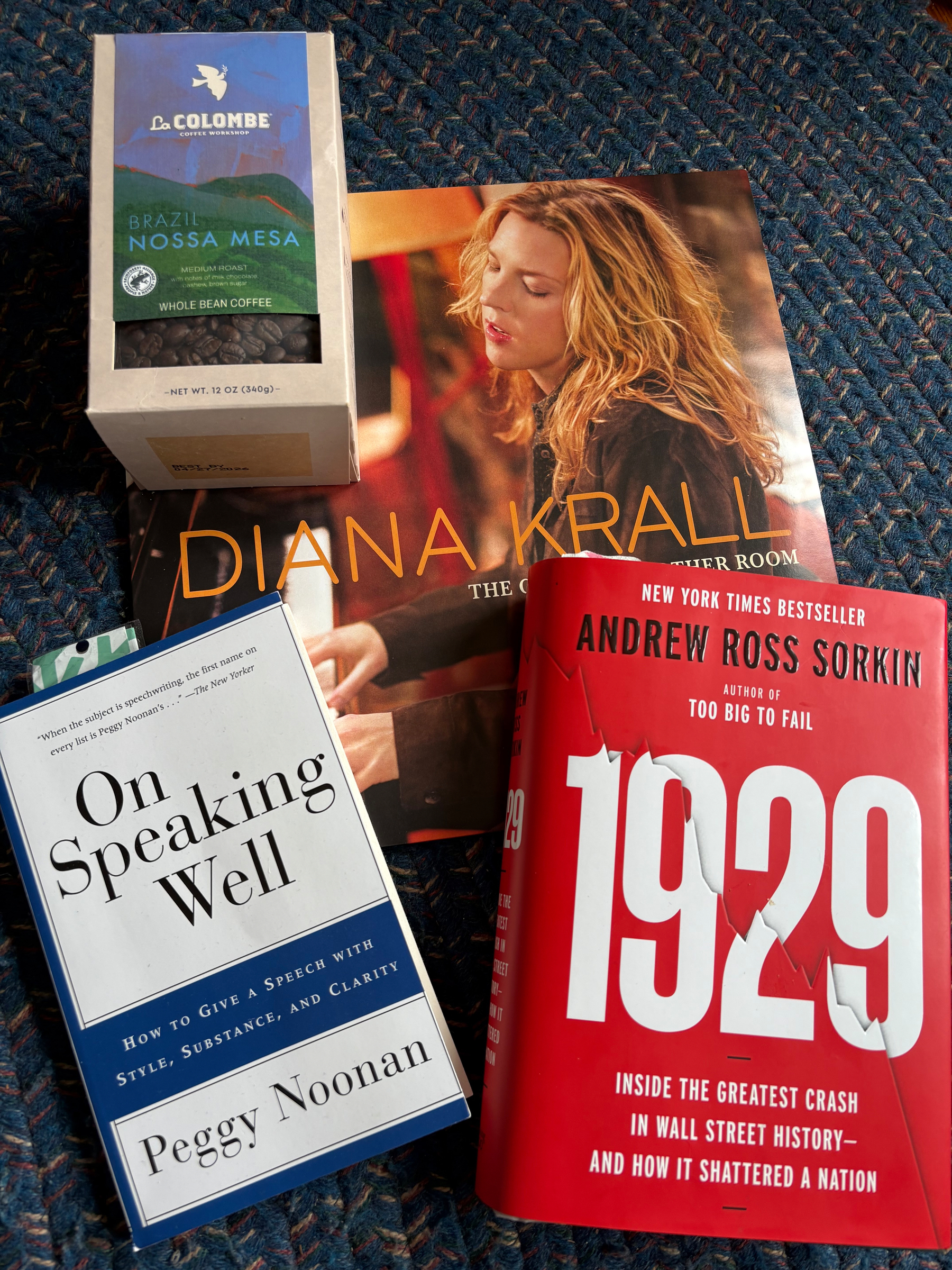 Auto-generated description: Coffee and books are arranged on a patterned surface, featuring titles by Diana Krall, Peggy Noonan, and Andrew Ross Sorkin.